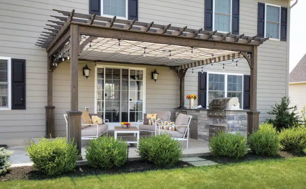 A backyard patio with a pergola, outdoor seating, string lights, and a built-in grill beside a house.
