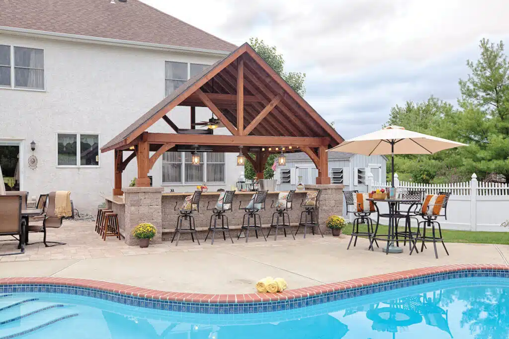 Outdoor patio with a wooden pergola bar, barstools, dining table, and pool beside a white house.