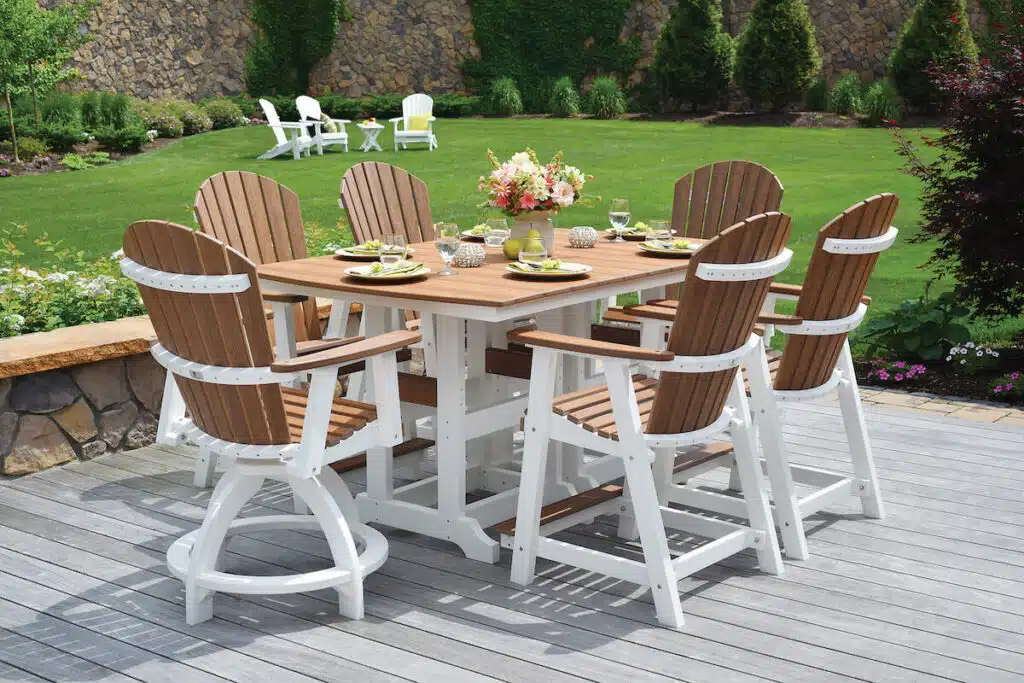 A wooden patio dining set with six chairs and a flower centerpiece, on a deck in a green backyard.