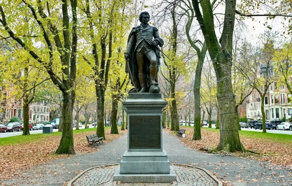 Bronze statue of a man on a stone pedestal in a tree-lined park with fallen autumn leaves.