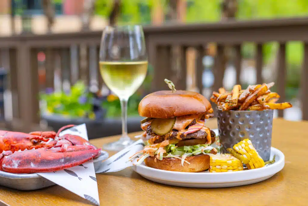 Burger with fries, corn, lobster, and a glass of white wine on an outdoor table.