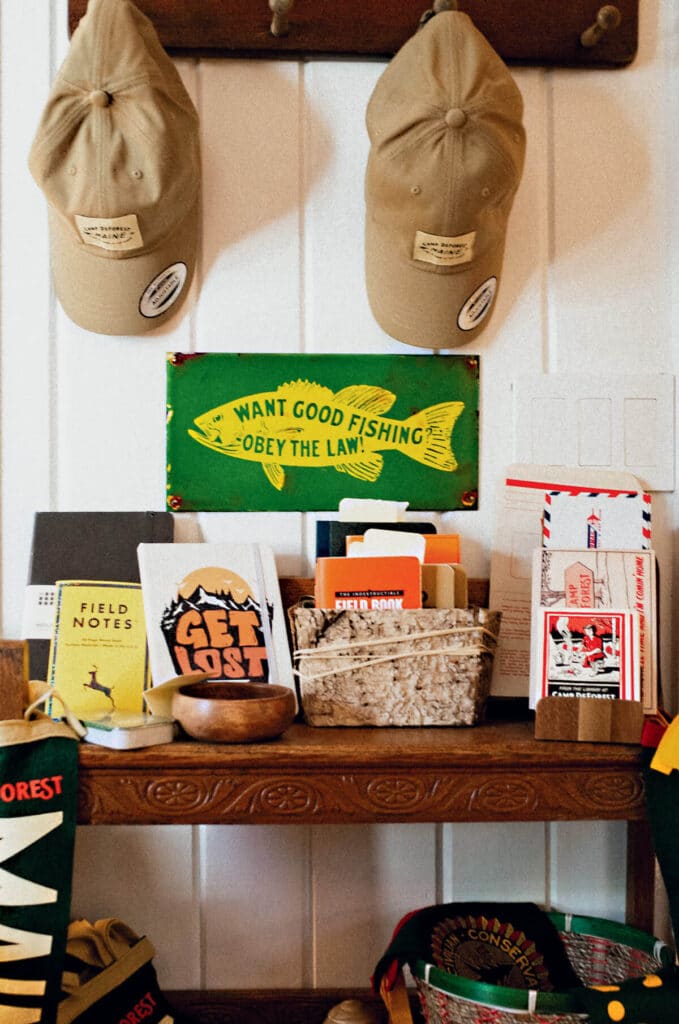 Two tan caps hang above a shelf with books, baskets, and a green fishing sign reading "Want Good Fishing Obey The Law.