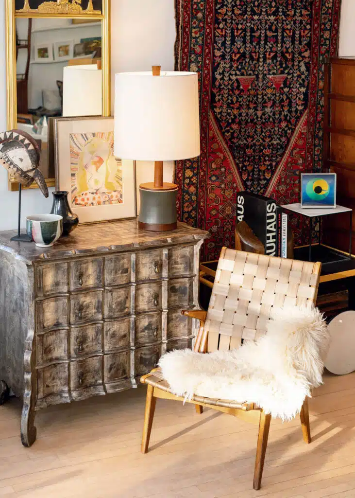 Eclectic room with a woven chair, fur throw, rustic chest, art, lamp, and patterned red rug.