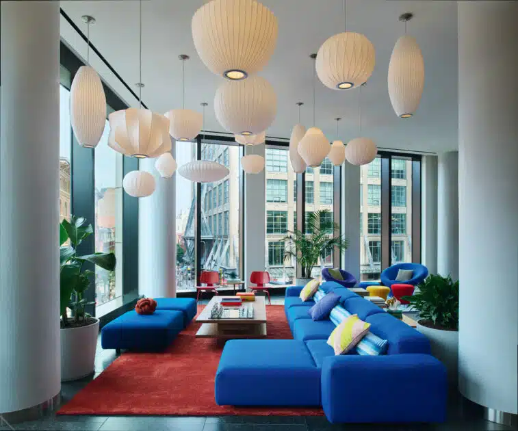 Modern lounge with blue sofas, colorful pillows, red chairs, large windows, plants, and white pendant lights.