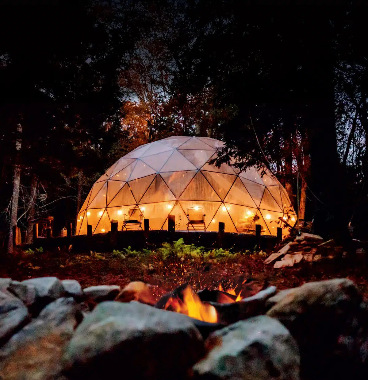 A glowing geodesic dome at night in a forest, with a campfire burning in the foreground.