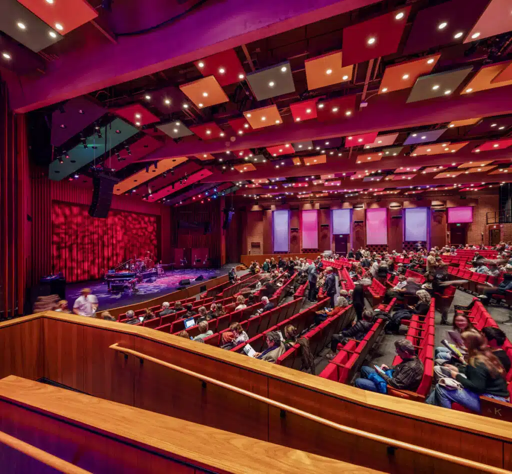Theater with red seats, colorful ceiling panels, and audience members waiting for a show to start.