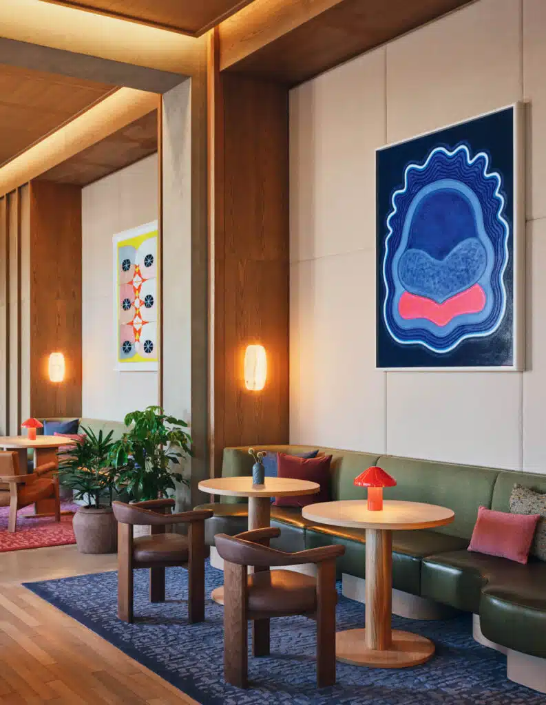 Modern lounge with wooden tables, green bench seating, colorful art, and warm wall lighting.