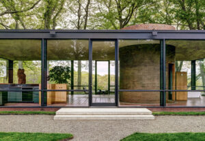 Modern glass house with floor-to-ceiling windows, brick cylinder inside, surrounded by trees and greenery.