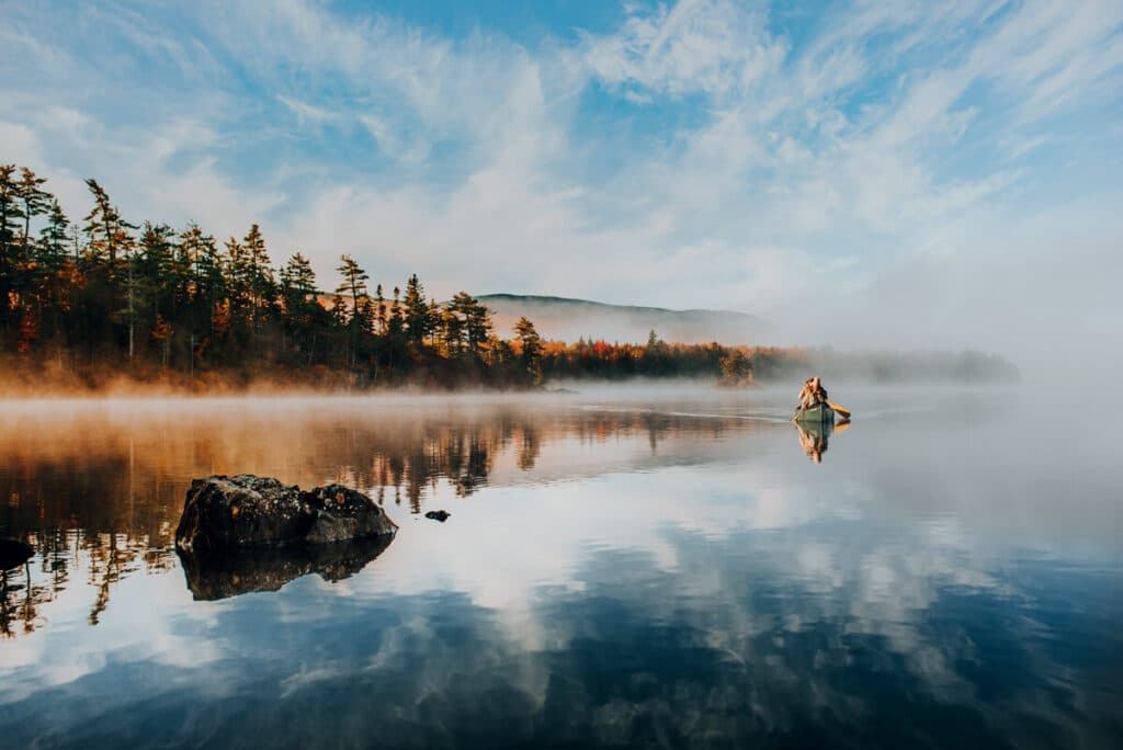 A canoe floats on a calm, misty lake surrounded by trees with autumn colors and reflections in the water.