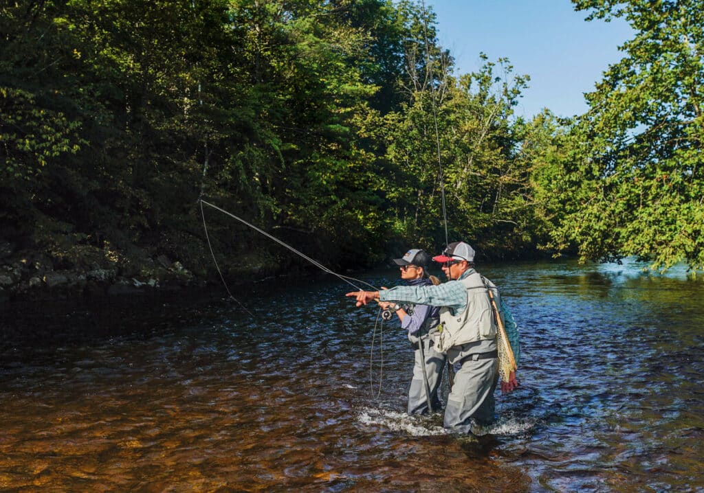 Two people fly fishing in a shallow, sunlit river surrounded by green trees, one pointing ahead.