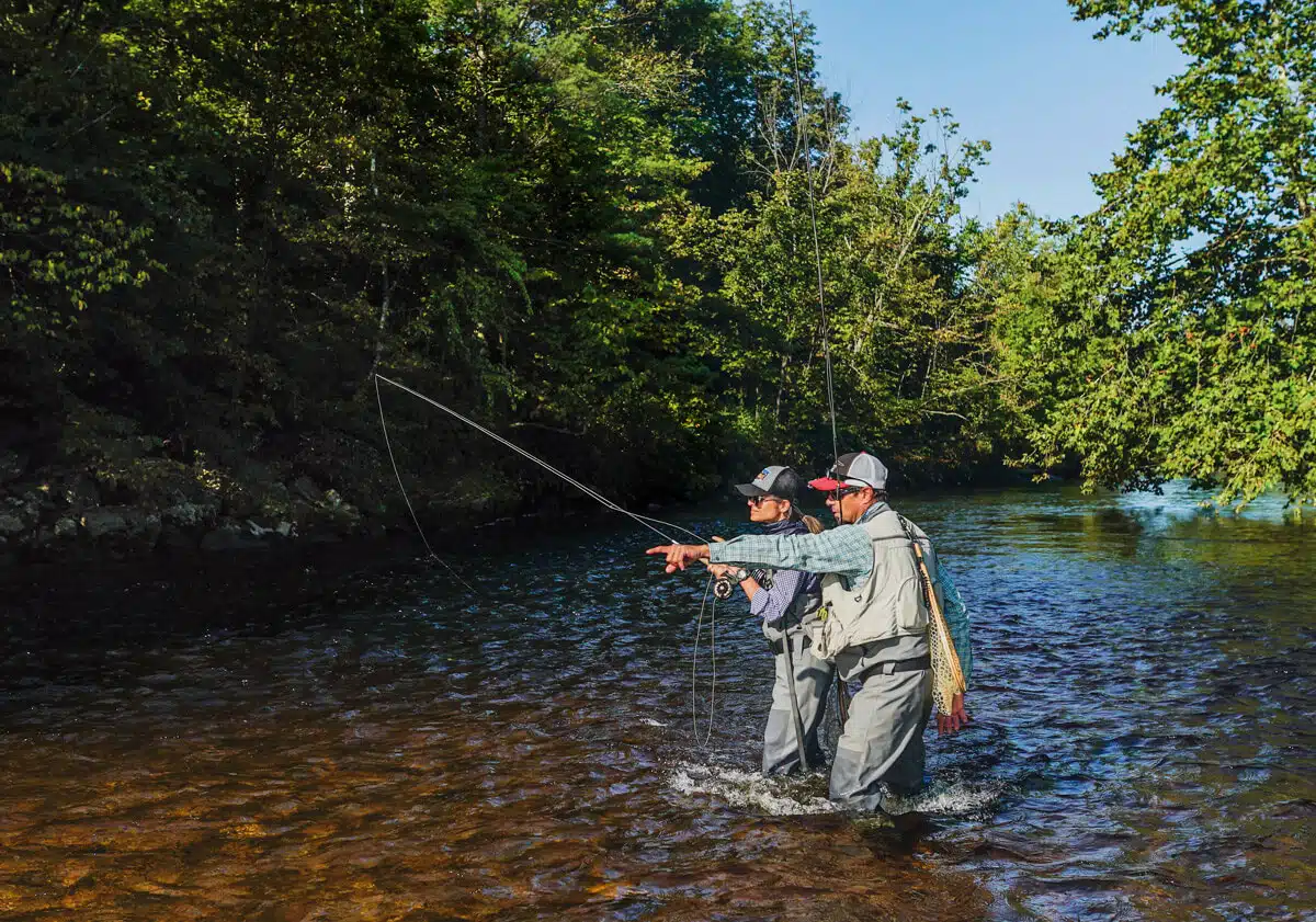 Two people fly fishing in a shallow, sunlit river surrounded by green trees, one pointing ahead.