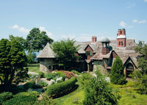 2026 Massachusetts Travel Guide. Large stone house with wooden roof, lush garden, chimney, and trees under a clear blue sky.