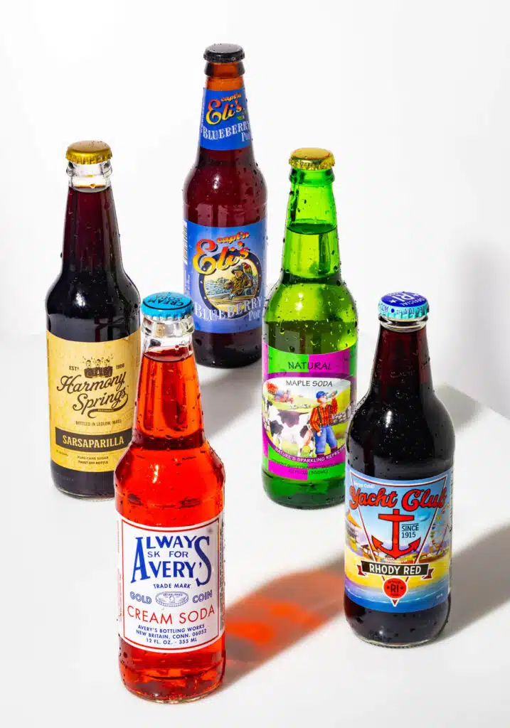 Pop Stars: New England’s Craft Soda Standouts. Five colorful bottles of craft soda arranged upright on a white surface, each with distinct labels.