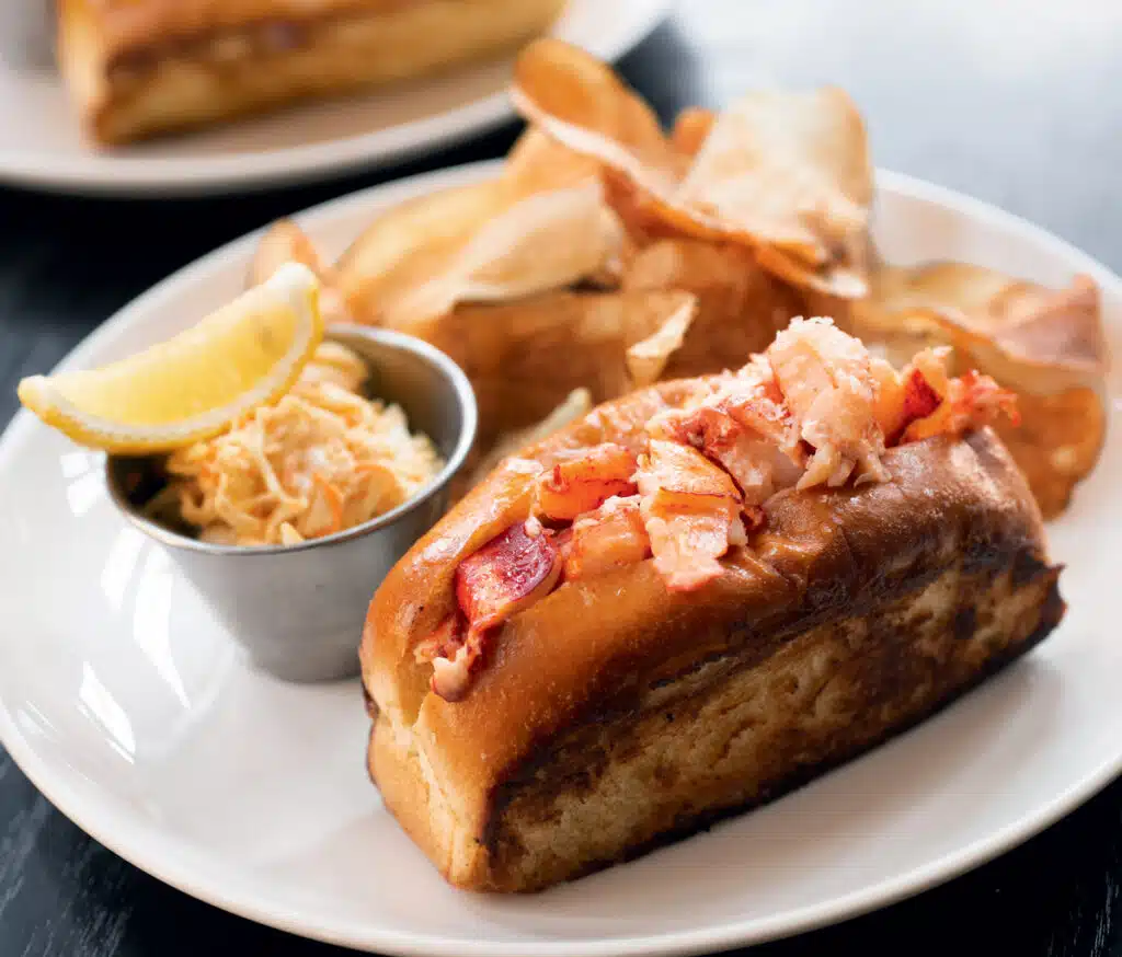 A lobster roll with potato chips and coleslaw, served on a white plate with a lemon wedge.
