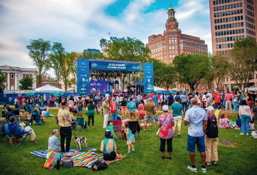 2026 Connecticut Travel Guide | Hotels, Dining & Attractions. Crowd gathered on a grassy lawn watching a live performance on an outdoor stage in a city park.