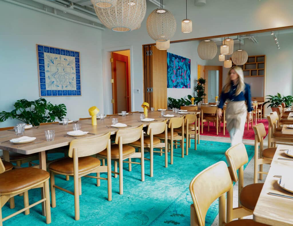 Modern restaurant with wooden furniture, blue rug, hanging lamps, and a woman walking past set tables.