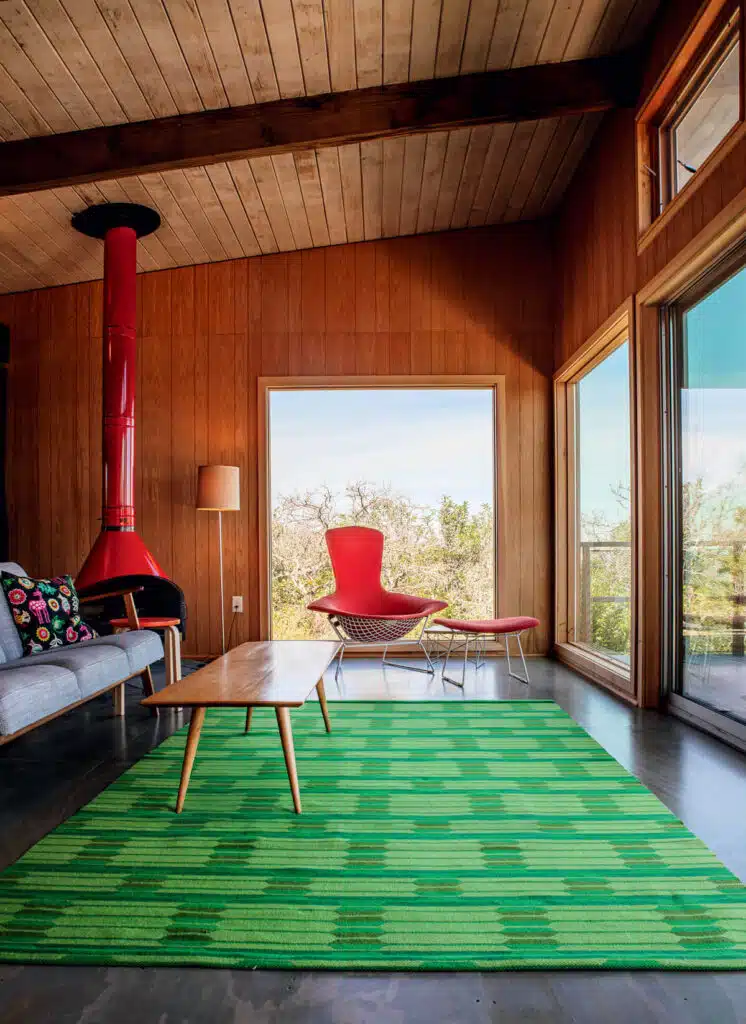 Finding Midcentury Modern in New England. Mid-century modern living room with wood walls, green rug, red chair, and red fireplace by large windows.
