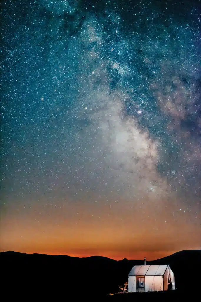 True North: Wild Landscapes From Cait Bourgault. A tent glows under a star-filled night sky with the Milky Way visible above dark silhouettes of hills.