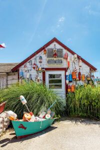 Small rustic shack decorated with colorful buoys, tall grass, and a boat in front under a bright blue sky.