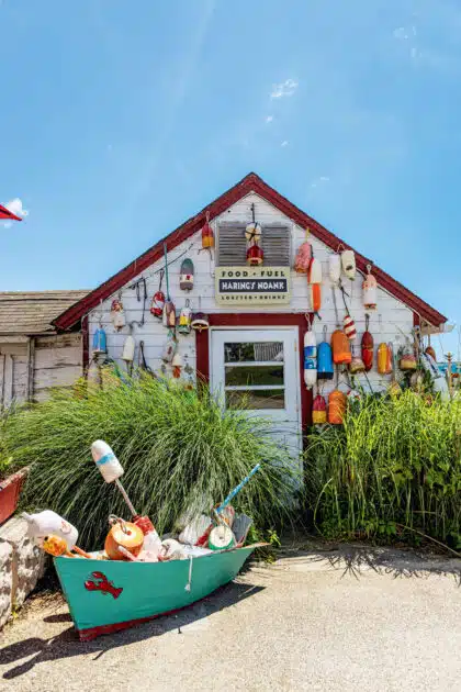 Small rustic shack decorated with colorful buoys, tall grass, and a boat in front under a bright blue sky.
