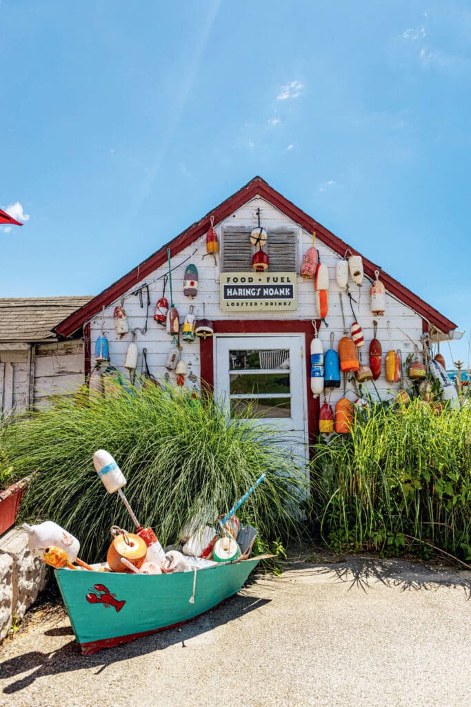 Small rustic shack decorated with colorful buoys, tall grass, and a boat in front under a bright blue sky.