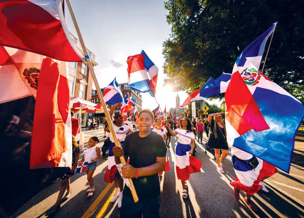 2026 Massachusetts Travel Guide | Hotels, Dining & Attractions. Children and adults march in a parade, waving Dominican Republic flags on a sunny street.