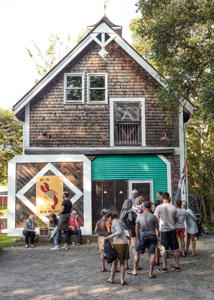 People stand in line outside a rustic building with a lobster sign and shingled exterior on a sunny day.