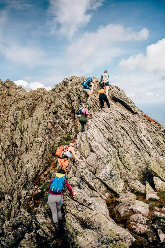 Five hikers with backpacks climb a rugged, rocky mountain ridge under a partly cloudy sky.