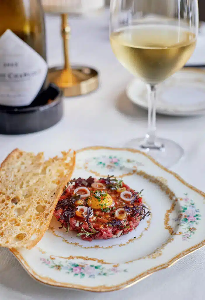 A fancy plate with steak tartare, herbs, a slice of toasted bread, and a glass of white wine on a table.