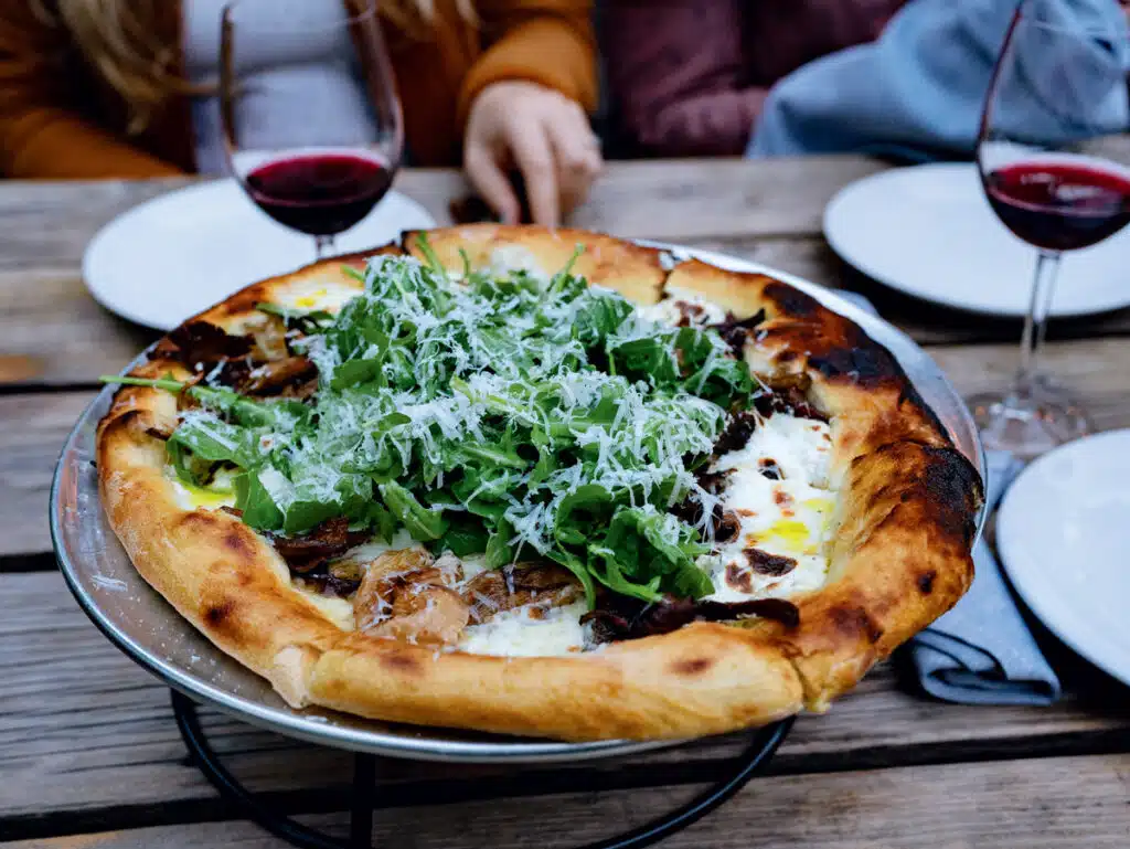 A pizza topped with arugula and cheese sits on a stand, with two glasses of red wine and plates nearby.