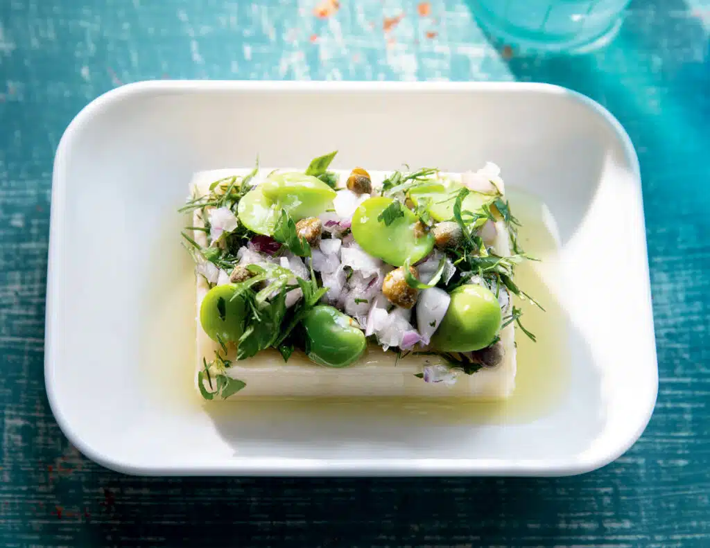 A rectangular block of cheese topped with broad beans, herbs, onions, and capers in a white dish.