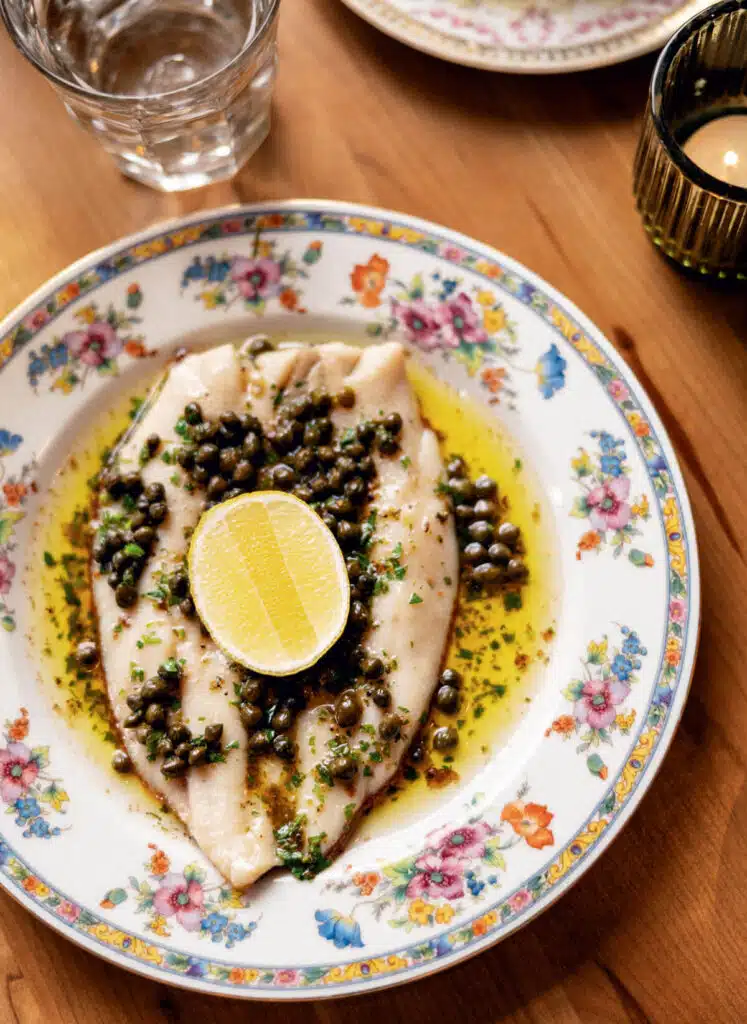 2026 Vermont Travel Guide | Hotels, Dining & Attractions. A fillet of fish with capers, herbs, and lemon on a floral plate, next to a glass of water and a candle.