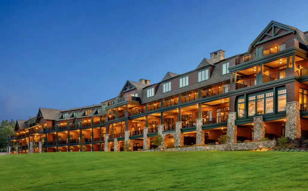 Large, multi-story lodge with stone pillars and warm lights, set against a green lawn and clear evening sky.
