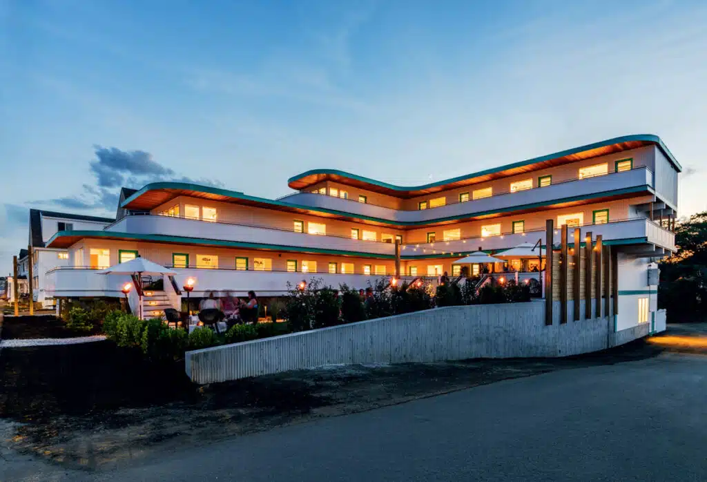 Modern, three-story building with glowing lights at dusk, surrounded by greenery and a curved driveway.