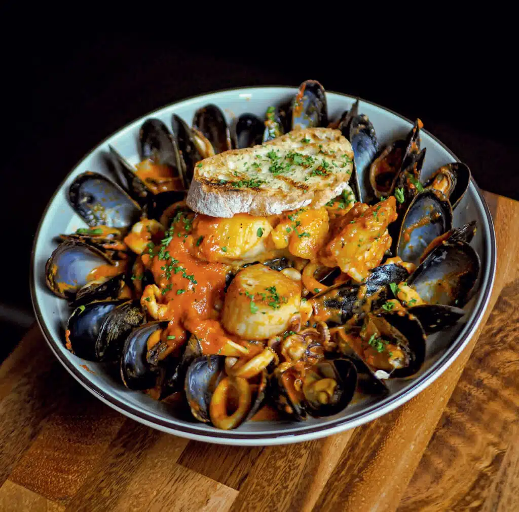 2026 Massachusetts Travel Guide | Hotels, Dining & Attractions. A bowl of mussels in tomato sauce, garnished with herbs and a slice of toasted bread on top.