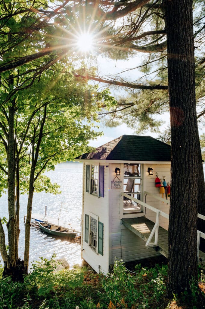 2026 Maine Travel Guide. Sunlight filters through trees onto a small lakeside cottage with a boat docked nearby.
