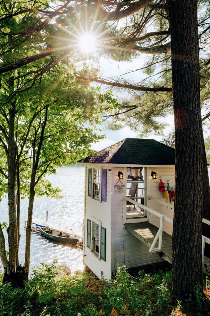 Sunlight filters through trees onto a small lakeside cottage with a boat docked nearby.