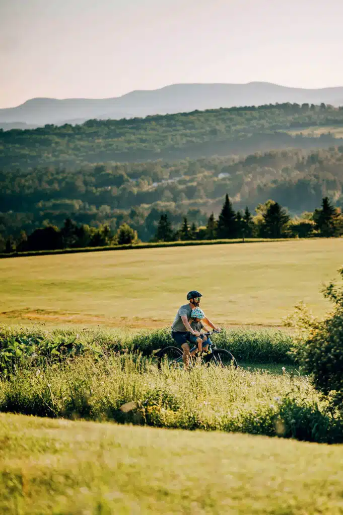 A person rides a bicycle with a child on a grassy path, with green hills and mountains in the background.