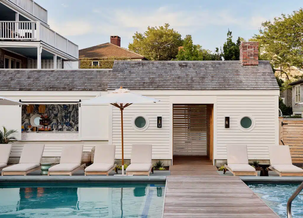 Poolside loungers and umbrellas beside a white pool house with round windows and wooden walkway.