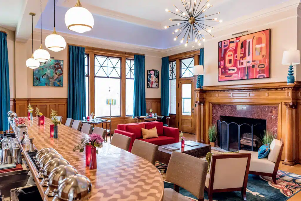 Colorful modern lounge with a wavy bar, red chairs, fireplace, large windows, and abstract art on the walls.