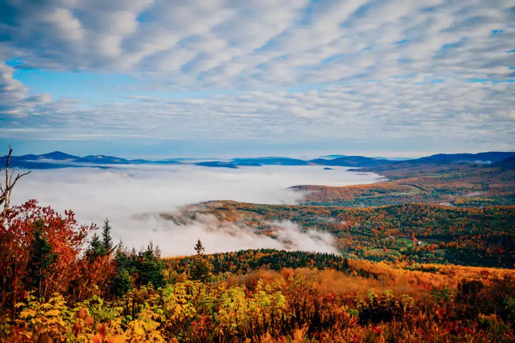 Colorful autumn forest landscape with rolling hills, low clouds, and a sky filled with scattered clouds.