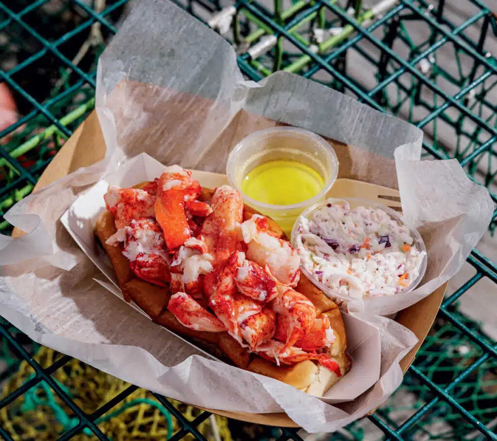 Lobster roll with a side of coleslaw and melted butter, served in a paper tray on a wire mesh table.