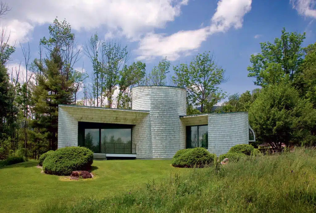 Modern house with circular and angular shapes, large windows, surrounded by green lawn, trees, and blue sky.