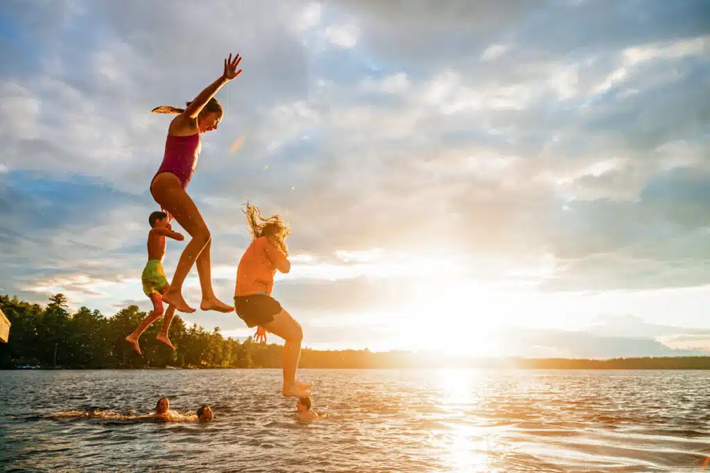 Three children joyfully jump into a lake at sunset, with others swimming in the water below.