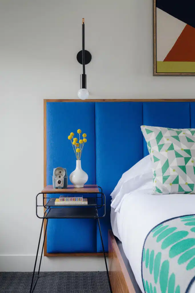 Nightstand with a vase of yellow flowers, retro clock, and modern blue upholstered headboard in a bright bedroom.