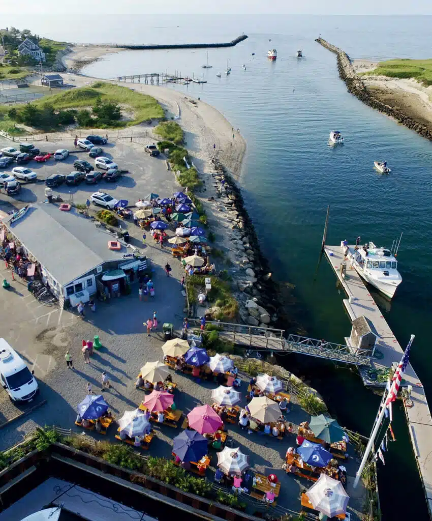 Aerial view of a waterfront restaurant with colorful umbrellas, parked cars, and boats on the water.