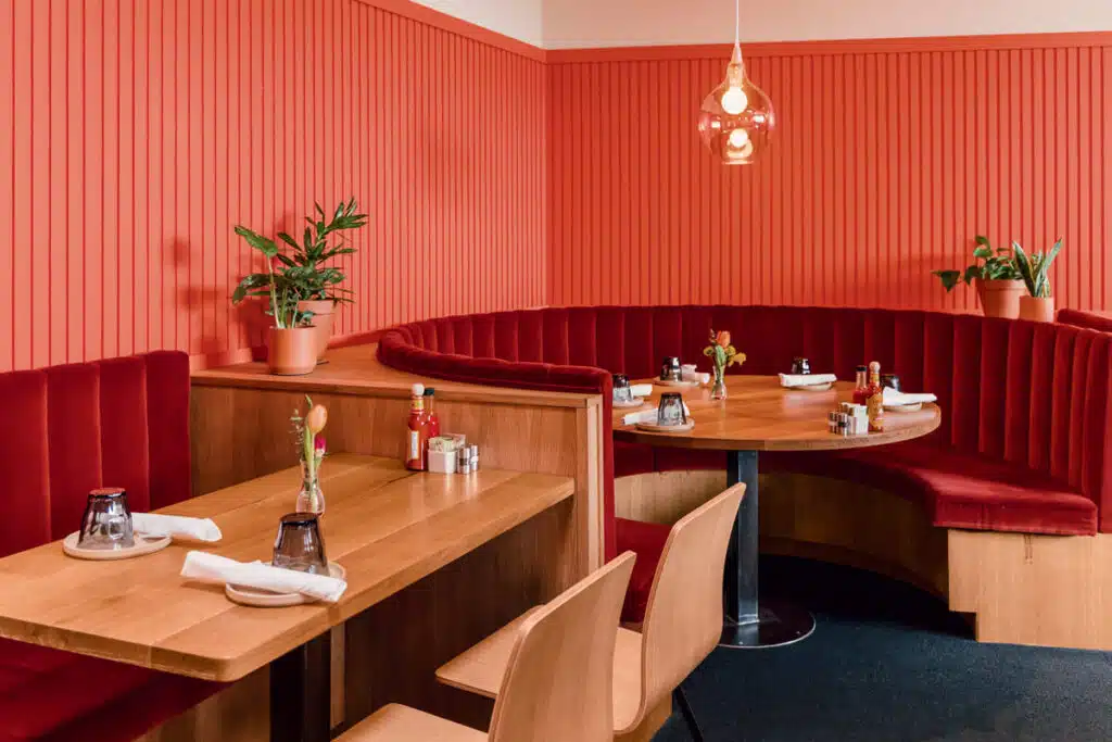 A modern restaurant with red velvet booths, wood tables, potted plants, and a hanging pendant light.