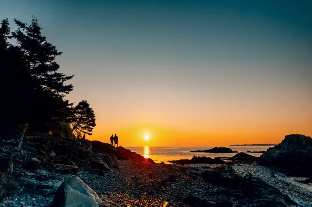 Two people stand on a rocky shore at sunset, with the sun low over the calm ocean and trees on the left.