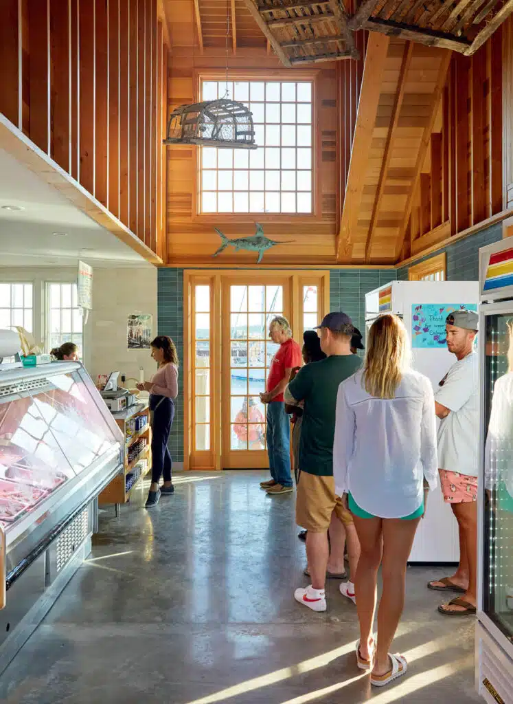 People stand in line at a bright, rustic seafood market with large windows and wooden beams.