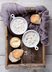 Two bowls of creamy soup with biscuits and butter on a rustic wooden tray lined with a purple cloth.